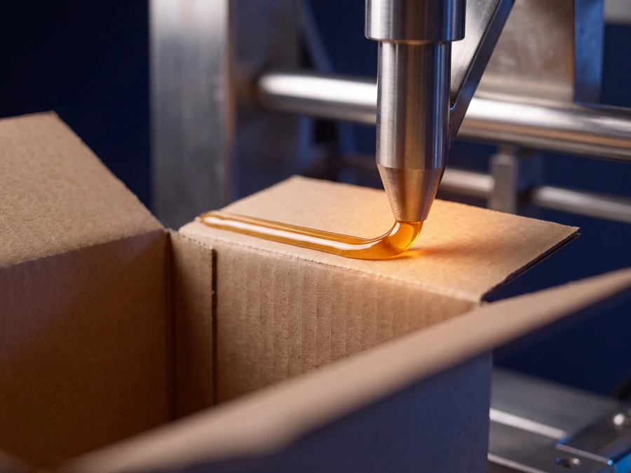 Close-up of a carton sealing line applying a bead of hot-melt adhesive to a corrugated flap